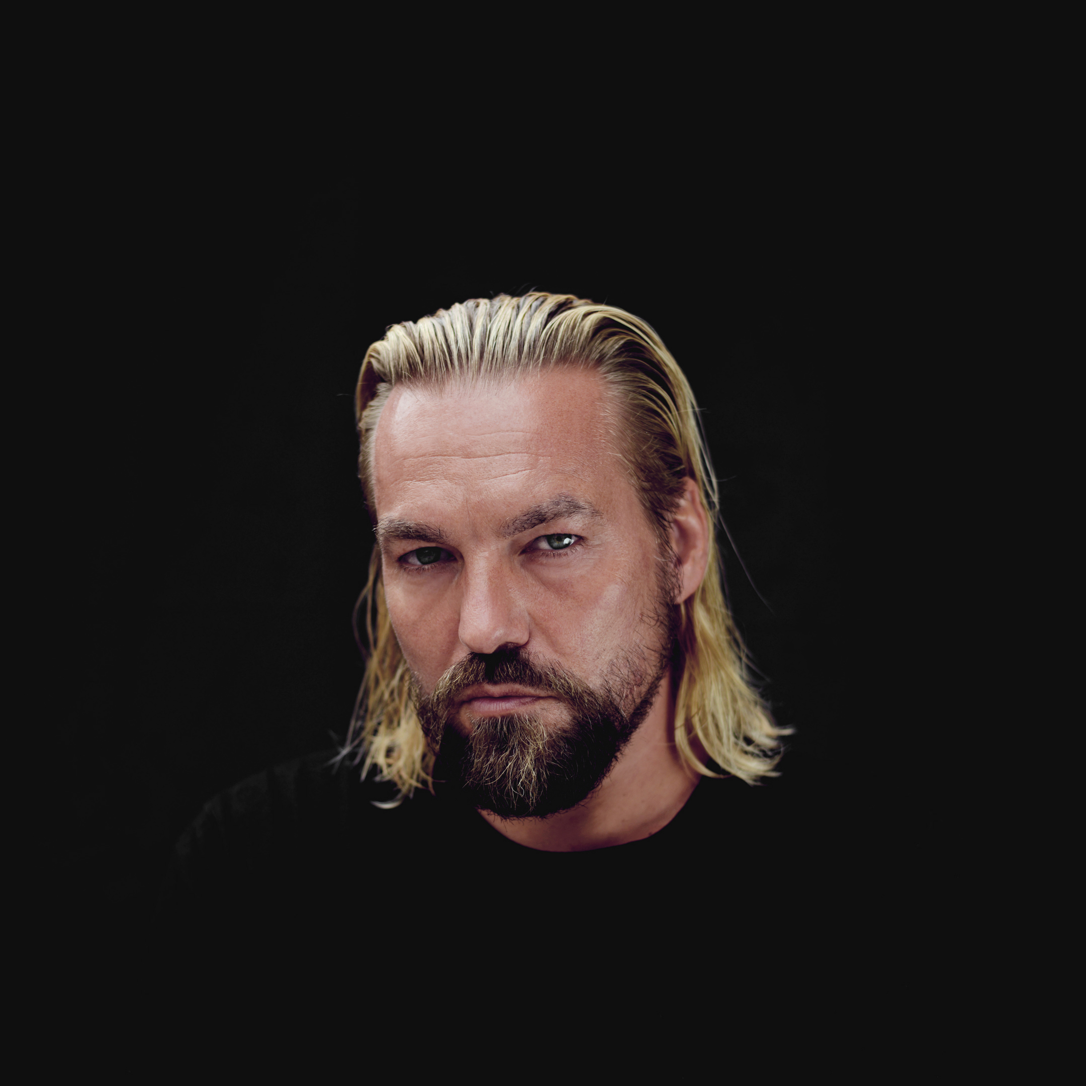 Portrait von Tai Lückerath vor schwarzem Hintergrund, mit blondem Haar und Bart, direkter Blick in die Kamera.